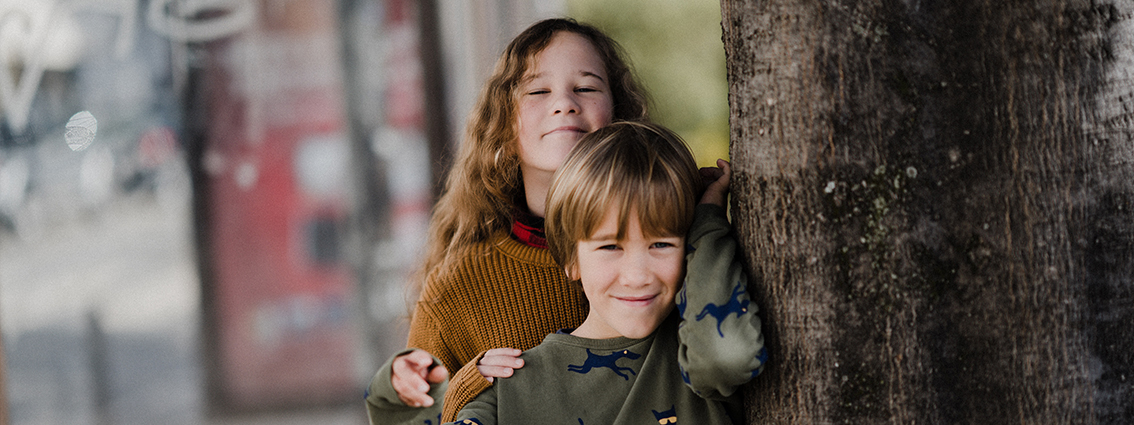 Foto eines Mädchens und eines kleinen Jungen, welche lächelnd hintereinander an einem Baum angelehnt stehen.